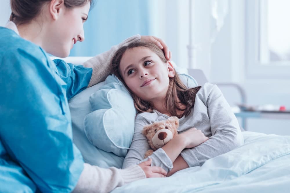Young girl in a hospital bed holding a teddy bear with her nurse stroking her head